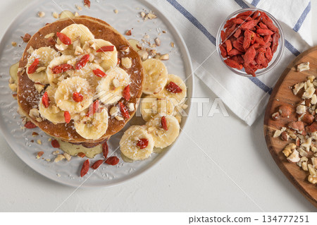 Pancakes with banana, walnuts, goji and honey. Healthy breakfast. On white wooden table background. Top view Pancakes with banana, walnuts, goji and honey. Healthy breakfast. On white wooden table background. Top view 134777251