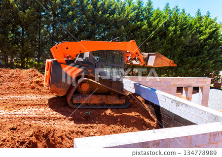 Small excavator moves dirt at construction site surrounded foundation of building 134778989