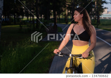 A Caucasian woman in yellow leggings and a crop top rides a bicycle in tights in the park.  134780682