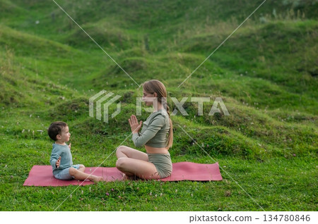 Caucasian woman doing yoga with her little son outdoors.  134780846
