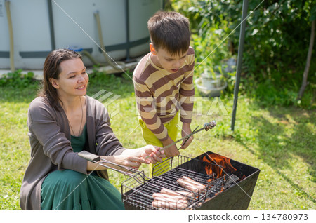 Caucasian woman grilling sausages on a barbecue with her two sons. Caucasian woman grilling sausages on a barbecue with her two sons. 134780973