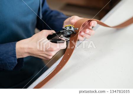 A woman makes holes in a leather belt. Leatherworker's workshop. A woman makes holes in a leather belt. Leatherworker's workshop. 134781035