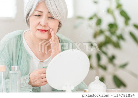 一位頭髮花白的老婦人在家中照鏡子，擔心自己的法令紋和嘴角。 134781854