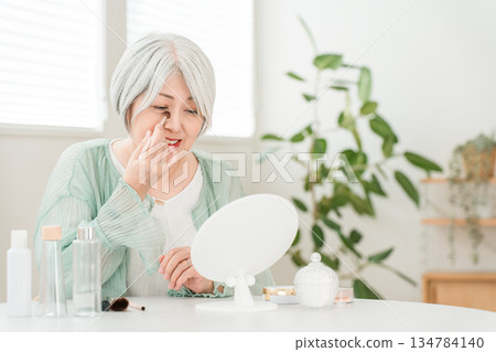 A senior woman with gray hair applying makeup and eye makeup while looking in the mirror at home 134784140