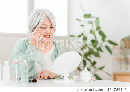 一位頭髮花白的老婦人正在家裡對著鏡子化妝，畫眼妝。 134784141