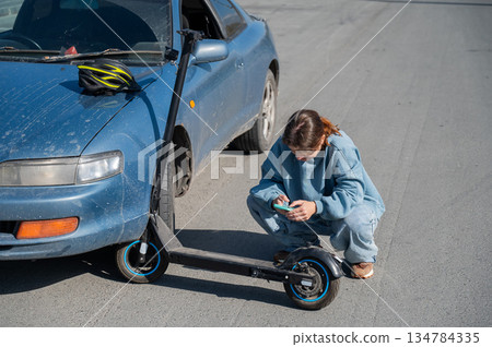 Woman examines and photographs the damage caused by a collision between an electric scooter and a car. Woman examines and photographs the damage caused by a collision between an electric scooter and a car. 134784335