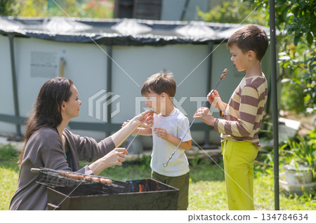 Caucasian woman grilling sausages on a barbecue with her two sons.  134784634