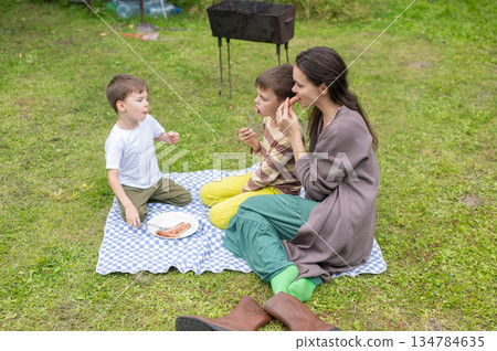 Caucasian woman eats fried sausages with her sons at a picnic. Caucasian woman eats fried sausages with her sons at a picnic. 134784635