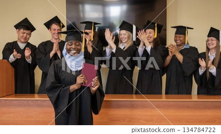 Happy African woman in hijab receiving diploma and classmates applauding her.  134784722