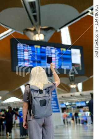 Passenger checks flight details at an airport terminal while holding a passport in the air and preparing for travel during a sunny day 134786915