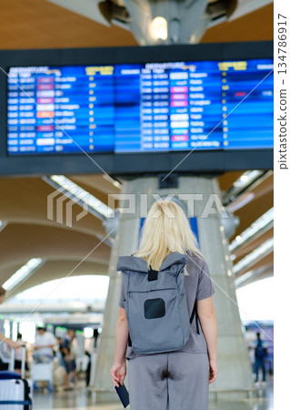 Traveler waiting at the airport terminal looking at the flight information board displaying departure and arrival times in a busy travel environment 134786917