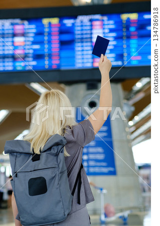 Traveler with a backpack raises a passport at the airport while checking flight information on a digital display board Traveler with a backpack raises a passport at the airport while checking flight information on a digital display board 134786918
