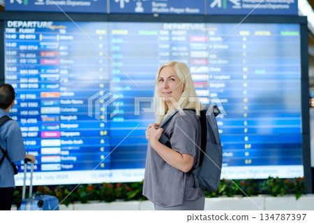 Woman with blonde hair stands near departure board at an airport, ready for an upcoming flight while travellling with a backpack on a sunny day Woman with blonde hair stands near departure board at an airport, ready for an upcoming flight while travellling with a backpack on a sunny day 134787397
