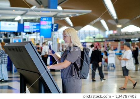 Woman checks in at an airport self-service kiosk while traveling, with busy terminal in the background during afternoon hours Woman checks in at an airport self-service kiosk while traveling, with busy terminal in the background during afternoon hours 134787402