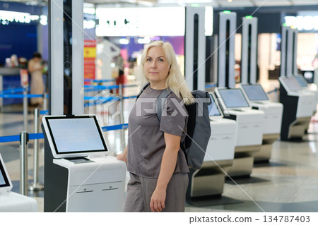 Woman with backpack interacting with check-in kiosk in modern airport terminal during busy travel season Woman with backpack interacting with check-in kiosk in modern airport terminal during busy travel season 134787403