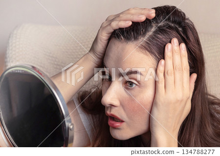 Young brunette woman, looks in the mirror at the first gray hair on her head. She's in shock. Copy space photo Young brunette woman, looks in the mirror at the first gray hair on her head. She's in shock. Copy space photo 134788277