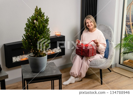Cozy holiday scene with woman sitting in chair and holding wrapped present indoors. Cozy holiday scene with woman sitting in chair and holding wrapped present indoors. 134788946