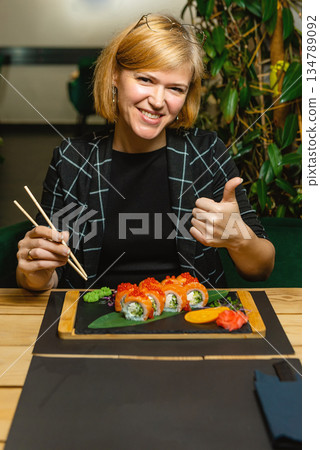 Smiling woman gives a thumbs-up while preparing to eat fresh sushi rolls. Smiling woman gives a thumbs-up while preparing to eat fresh sushi rolls. 134789092
