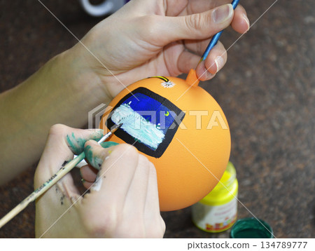 Female hands carefully painting a Christmas ball in vibrant orange hues Female hands carefully painting a Christmas ball in vibrant orange hues 134789777