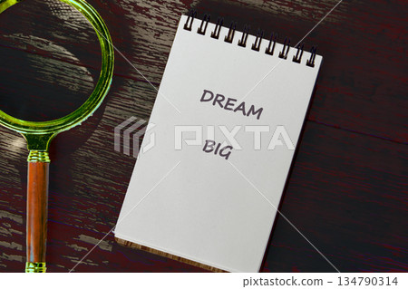 An open notebook with DREAM BIG inscribed lies beside a classic magnifying glass on a darkly stained wooden desk, alluding to grand ambitions and careful planning An open notebook with DREAM BIG inscribed lies beside a classic magnifying glass on a darkly stained wooden desk, alluding to grand ambitions and careful planning 134790314