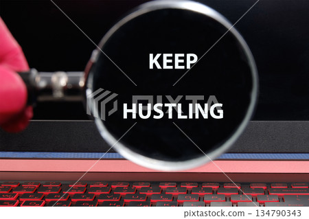 A pink-gloved hand holds a magnifying glass over a computer screen displaying the message KEEP HUSTLING. A red-lit keyboard sits in the foreground on the laptop A pink-gloved hand holds a magnifying glass over a computer screen displaying the message KEEP HUSTLING. A red-lit keyboard sits in the foreground on the laptop 134790343