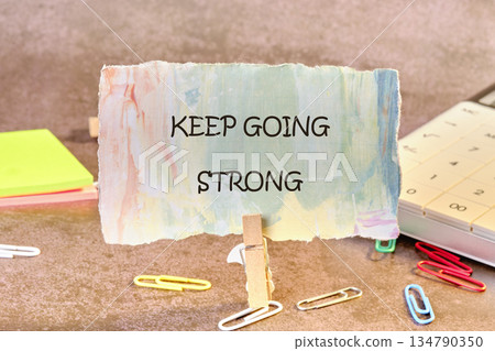 A small, colorful note with the message Keep Going Strong sits upright with a clothespin. Paperclips and notepads add a touch of creativity to the scene. A calendar sits nearby A small, colorful note with the message Keep Going Strong sits upright with a clothespin. Paperclips and notepads add a touch of creativity to the scene. A calendar sits nearby 134790350
