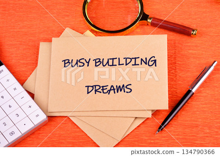 On an orange desk sits paper stating BUSY BUILDING DREAMS, alongside a calculator, magnifying glass, and pen. Tools for creating a brighter future On an orange desk sits paper stating BUSY BUILDING DREAMS, alongside a calculator, magnifying glass, and pen. Tools for creating a brighter future 134790366