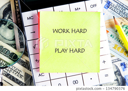 A yellow sticky note that reads work hard play hard sits on top of a white calculator surrounded by American money and a pencil. A magnifying glass rests on the cash A yellow sticky note that reads work hard play hard sits on top of a white calculator surrounded by American money and a pencil. A magnifying glass rests on the cash 134790376