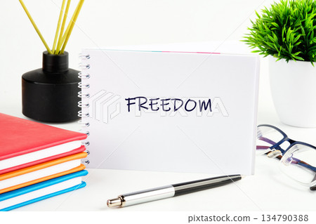 A white notebook with the word Freedom written on it rests on a desk. Colorful books, a pen, eyeglasses, and a green plant adorn the space, creating a serene setting 134790388