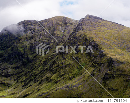 A scenic shot showcases the rugged terrain of the Coomloughra Horseshoe mountains. Green grass covers the slopes, with visible hiking trails. Clouds partially obscure the peaks. A scenic shot showcases the rugged terrain of the Coomloughra Horseshoe mountains. Green grass covers the slopes, with visible hiking trails. Clouds partially obscure the peaks. 134791504