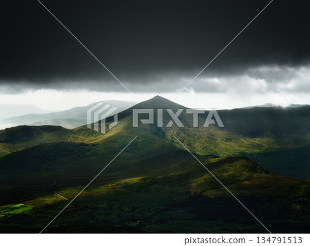 A stunning view of rugged mountain peaks in Ireland. Dark storm clouds loom above, while patches of sunlight illuminate the green valleys. 134791513