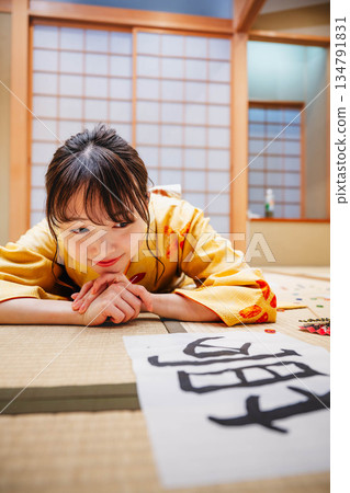 一位身著和服的年輕女子躺在日式房間裡,地板上散落著她的書法作品。 一位身著和服的年輕女子躺在日式房間裡,地板上散落著她的書法作品。 134791831