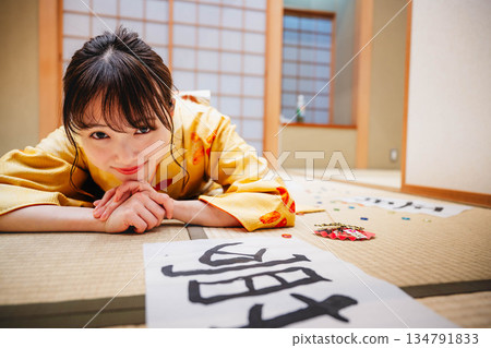 一位身著和服的年輕女子躺在日式房間裡,地板上散落著她的書法作品。 一位身著和服的年輕女子躺在日式房間裡,地板上散落著她的書法作品。 134791833