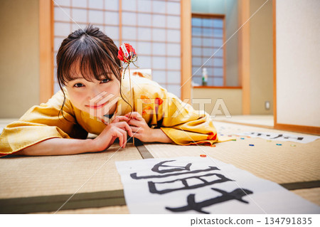 一位身著和服的年輕女子躺在日式房間裡，地板上散落著她的書法作品。 134791835