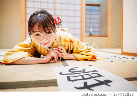 一位身著和服的年輕女子躺在日式房間裡，地板上散落著她的書法作品。 134791836