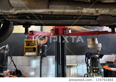 A car lift is positioned under a vehicle in an auto shop. Tools and equipment are visible in the area, indicating ongoing repairs and maintenance work 134792868