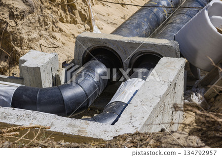 Heavy black pipes are connected to concrete blocks in an excavation site. Soil is moved aside, showing the depth of the work done on the drainage system Heavy black pipes are connected to concrete blocks in an excavation site. Soil is moved aside, showing the depth of the work done on the drainage system 134792957