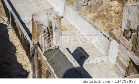 Workers prepare a concrete foundation with wooden forms on a construction site in daylight. The ground is uneven and dusty, showing raw materials and tools 134792958