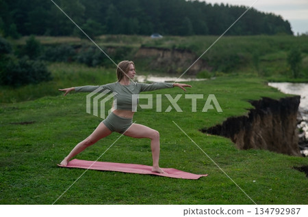 Caucasian woman doing yoga on the river bank. Warrior pose. Caucasian woman doing yoga on the river bank. Warrior pose. 134792987