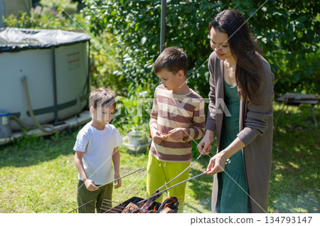 Caucasian woman grilling sausages on a barbecue with her two sons. Caucasian woman grilling sausages on a barbecue with her two sons. 134793147