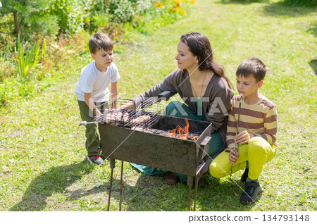Caucasian woman grilling sausages on a barbecue with her two sons.  134793148