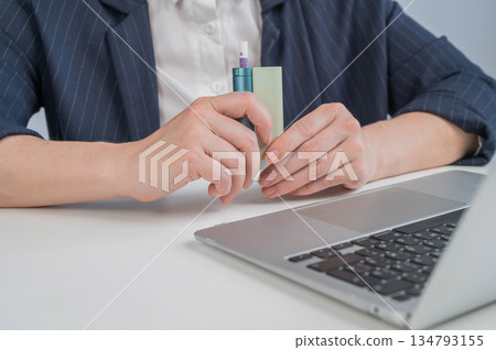 Business woman smoking an electronic cigarette while working on a laptop. Tobacco heating system. Business woman smoking an electronic cigarette while working on a laptop. Tobacco heating system. 134793155