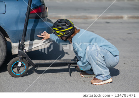 A distressed woman examines the damage after a collision between an electric scooter and a car. A distressed woman examines the damage after a collision between an electric scooter and a car. 134793471