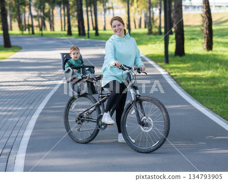 Caucasian woman riding a bicycle with her toddler son sitting behind her in a child seat.  134793905