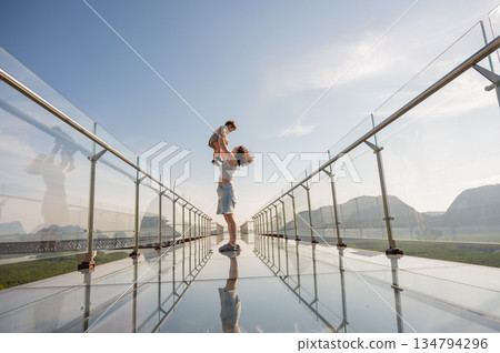 Caucasian woman holds her little son in her arms while standing on a glass bridge in the mountains. Caucasian woman holds her little son in her arms while standing on a glass bridge in the mountains. 134794296
