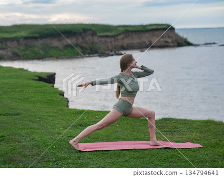 Caucasian woman doing yoga on the river bank. Warrior pose. Caucasian woman doing yoga on the river bank. Warrior pose. 134794641