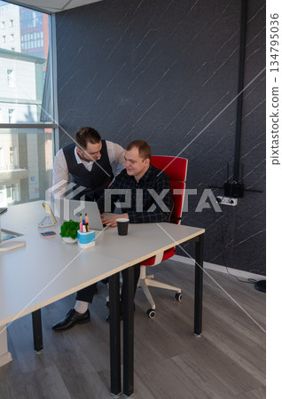 Caucasian man standing at colleague's desk discussing work.  134795036