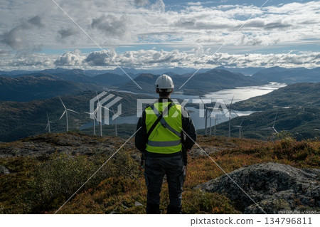 Technician in Neon Green Vest Observing Wind Farm Landscape with Mountains in Background Technician in Neon Green Vest Observing Wind Farm Landscape with Mountains in Background 134796811
