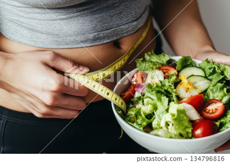Person Measuring Waistline with Tape While Holding Bowl of Fresh Vegetables Person Measuring Waistline with Tape While Holding Bowl of Fresh Vegetables 134796816
