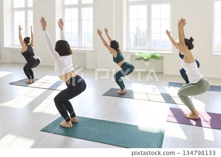 Yoga class practicing chair pose in bright studio with mats and morning light Yoga class practicing chair pose in bright studio with mats and morning light 134798133
